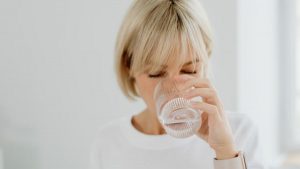 Blonde woman drinking water from glass indoors, showcasing healthy lifestyle and hydration.